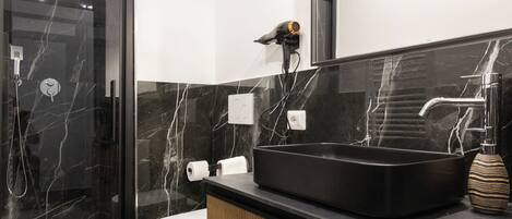A modern bathroom with a black sink, marble walls, a glass shower enclosure, and a wooden vanity.