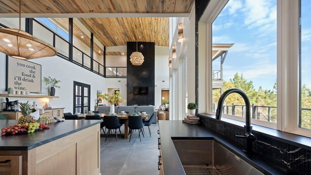 Una cocina moderna con una isla grande, encimeras negras y un lavamanos integrado. Hay una zona de comedor con mesa y sillas, y un ventanal que ofrece vistas al exterior.