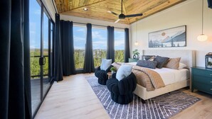 Un dormitorio con una cama grande, un ventilador de techo, una mesita de noche, un cuadro y vistas al exterior.