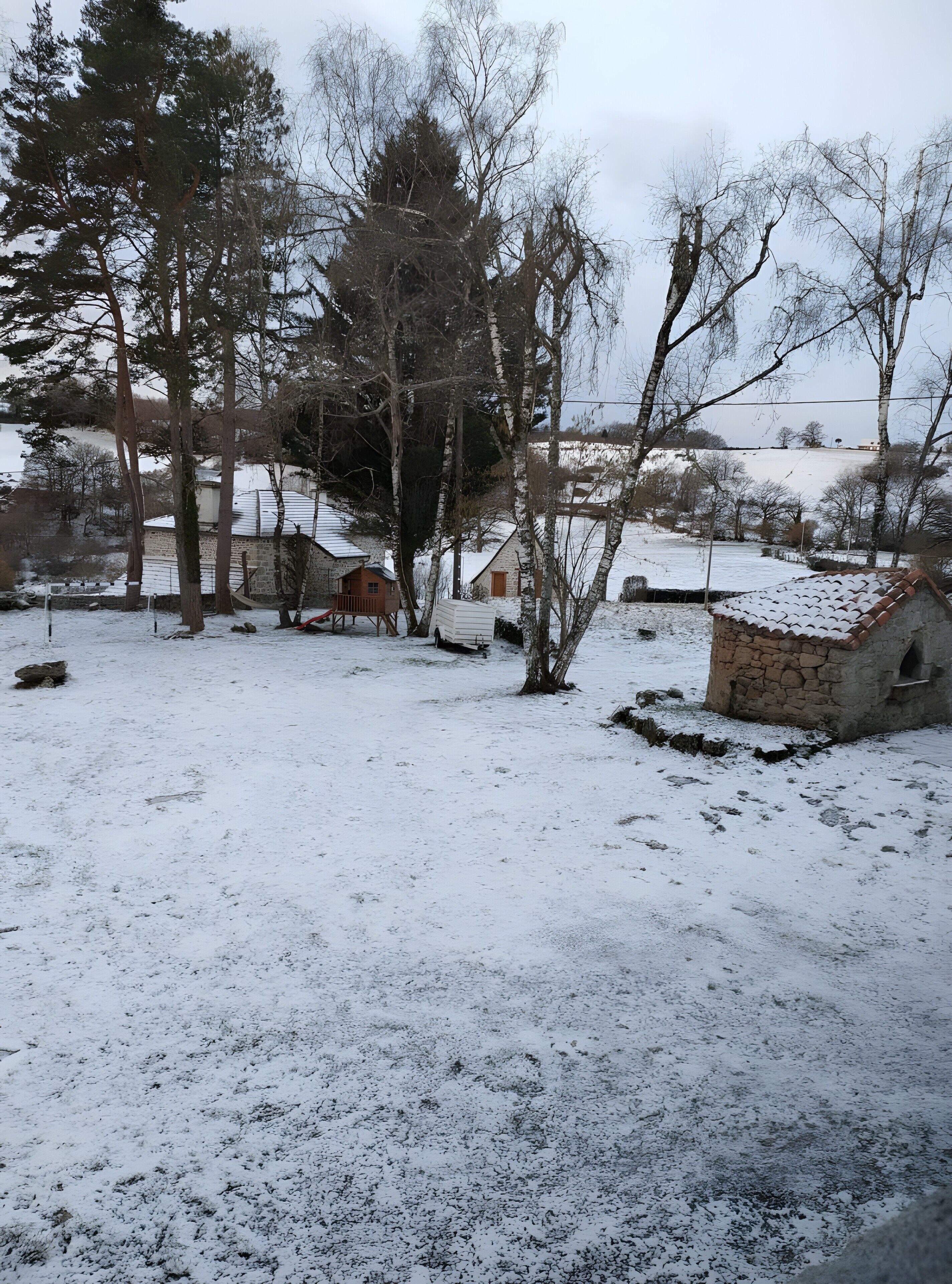 Un paysage enneigé avec un bâtiment en pierre, une structure en bois et une zone délimitée par une clôture.