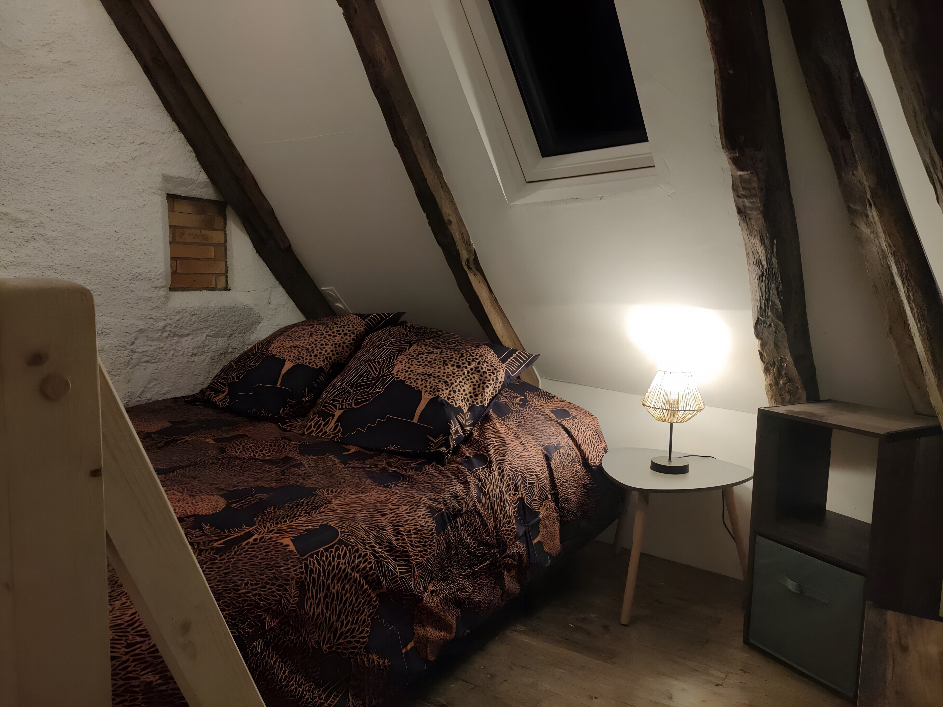 Et soveværelse med skrå væg, seng med mønstret sengetøj, natbord med lampe, og et lille træskab.