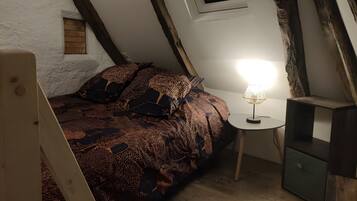 Et soveværelse med skrå væg, seng med mønstret sengetøj, natbord med lampe, og et lille træskab.