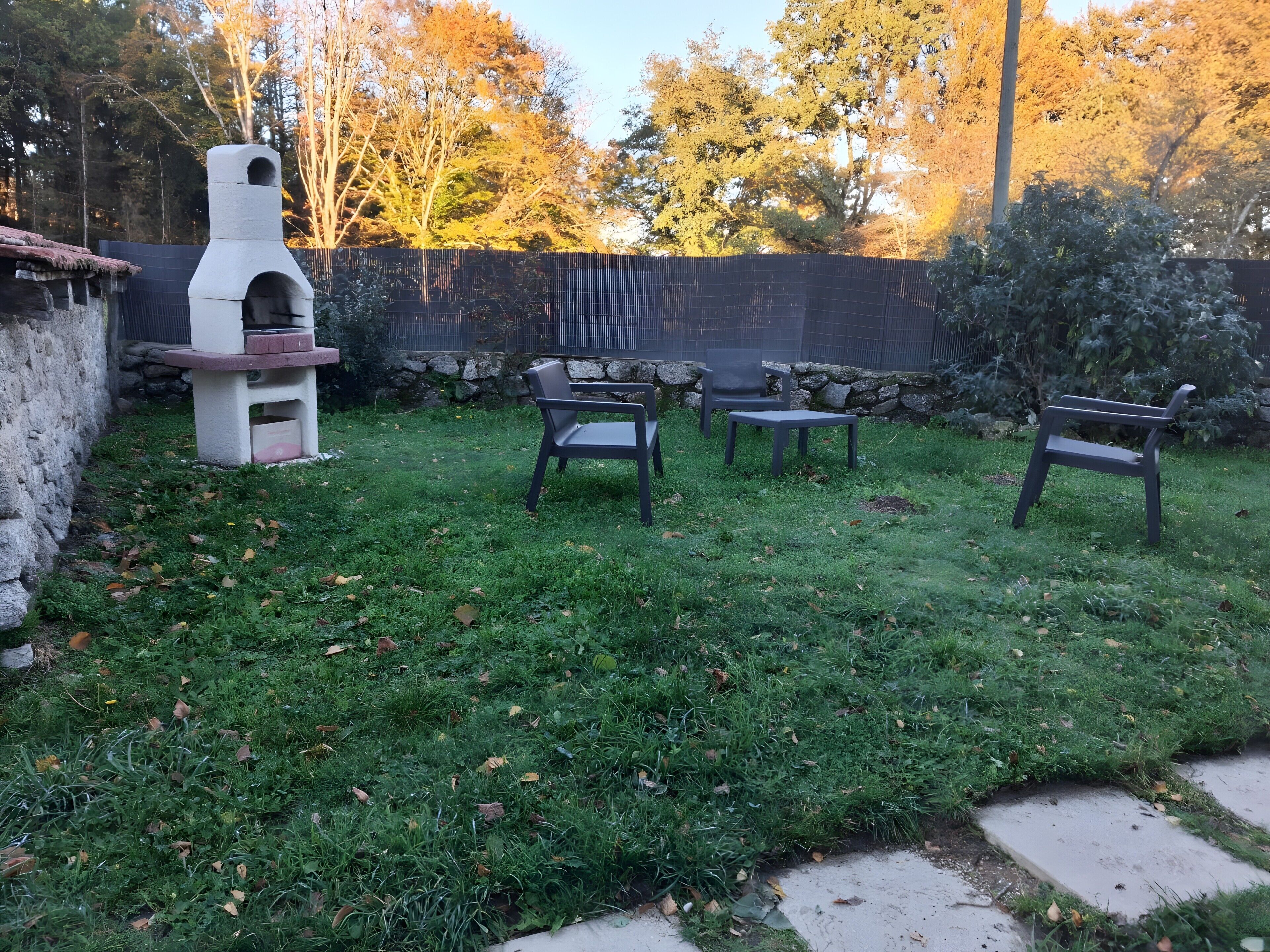 A backyard with a stone wall, a barbecue grill, and three chairs.
