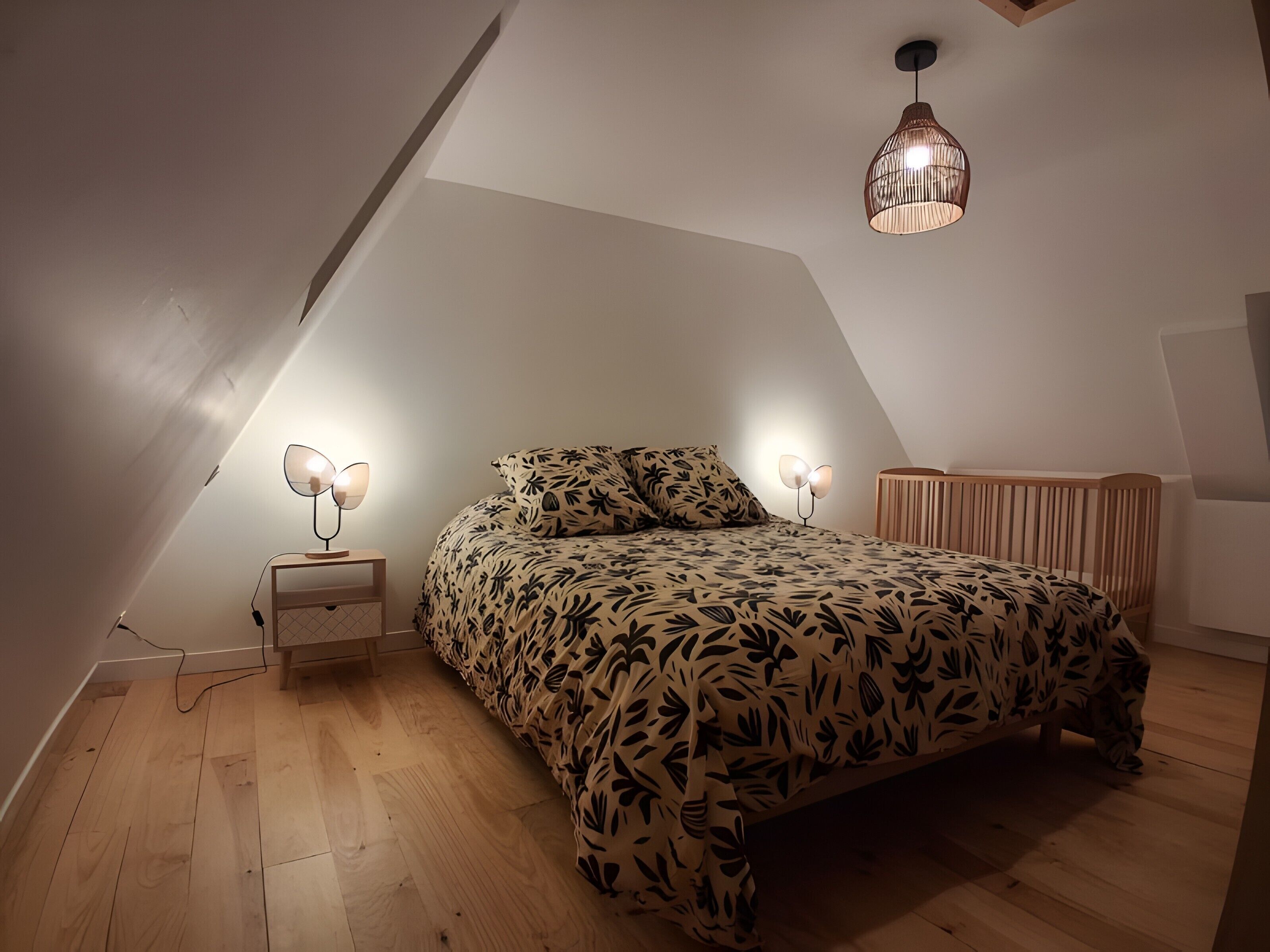 Un dormitorio con techo inclinado, una cama con colcha estampada con motivos de hojas, una lámpara de noche y una lámpara colgante.