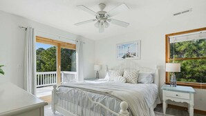 A bedroom with a bed, a nightstand, a ceiling fan, a window with a view of trees, and a door leading to an outdoor balcony. - Golf & Surf 7BR: Private Pool, 5 Min to Beach (Corolla)
