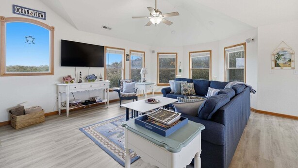 A spacious living room with a large window labeled "Ocean View," a ceiling fan, a flat-screen TV, a blue sofa, and a coffee table with magazines. - Golf & Surf 7BR: Private Pool, 5 Min to Beach (Corolla)