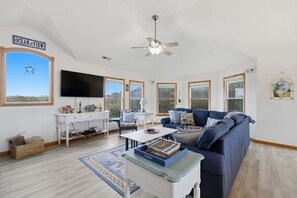 A spacious living room with a large window labeled "Ocean View," a ceiling fan, a flat-screen TV, a blue sofa, and a coffee table with magazines. - Golf & Surf 7BR: Private Pool, 5 Min to Beach (Corolla)