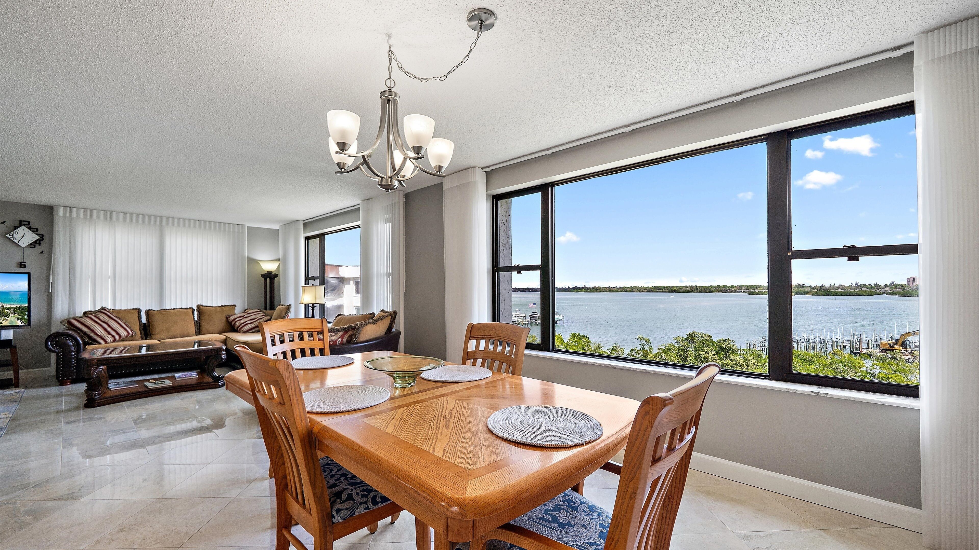 A spacious living room with a large dining table, comfortable seating, and a view of a body of water through large windows.