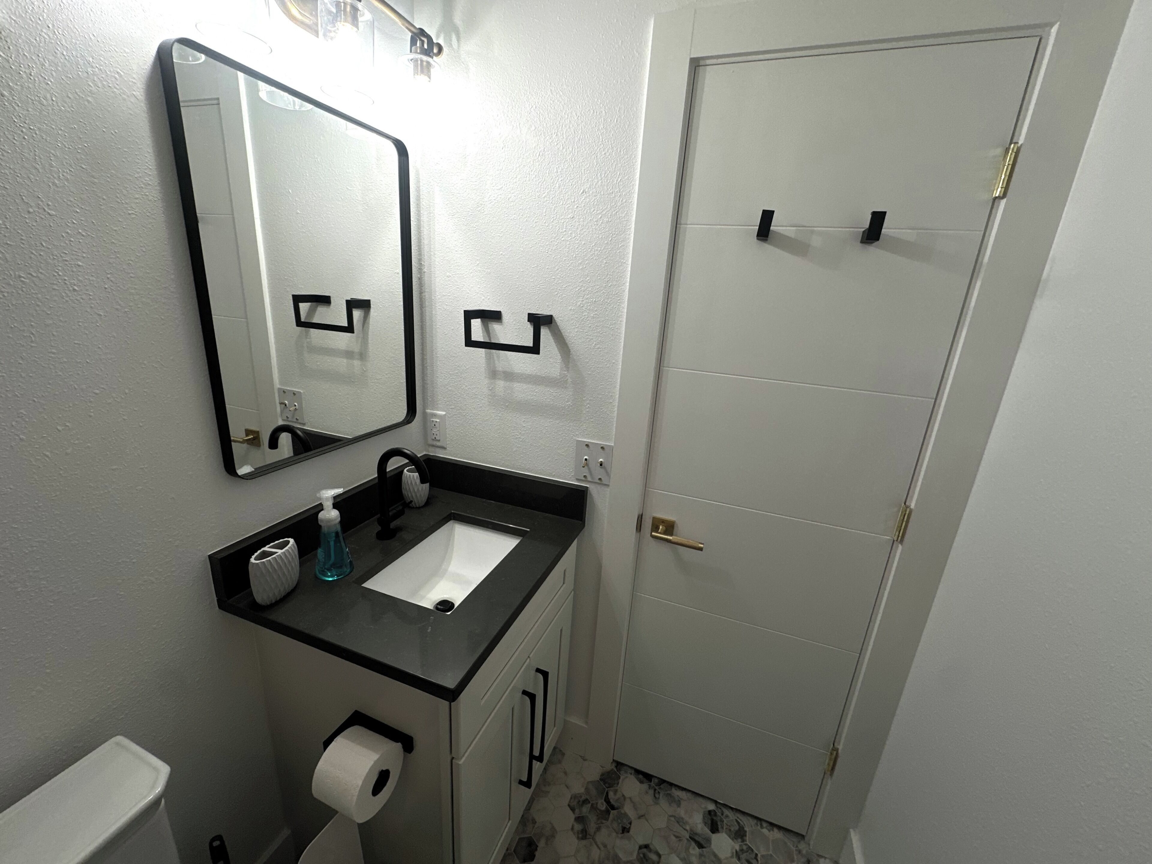 Une salle de bain avec un lavabo blanc, un plan de toilette noir et un miroir au-dessus. On y trouve des toilettes avec un rouleau de papier toilette et un flacon de savon pour les mains sur le plan de toilette. Le sol est recouvert de carreaux formant un motif.
