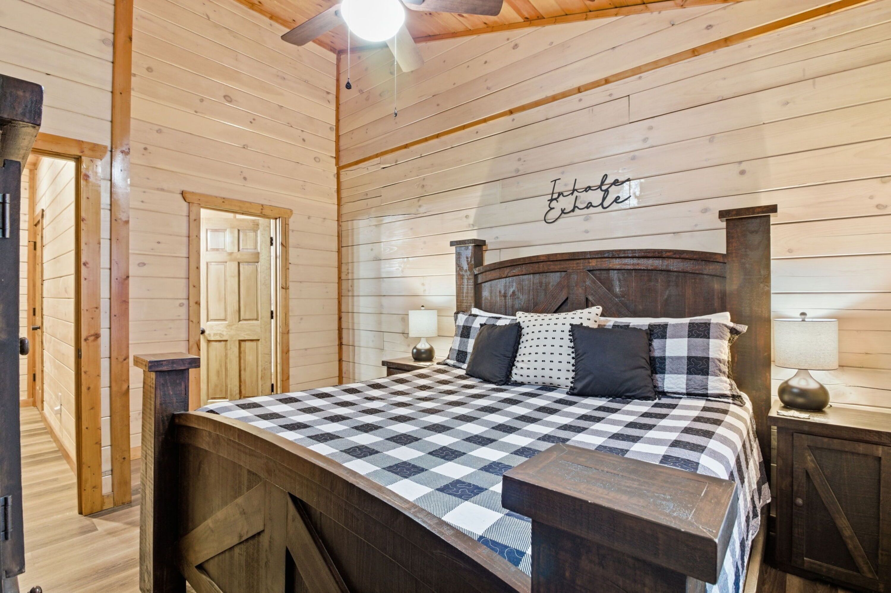 A wooden cabin bedroom with a checkered bedspread, a wooden headboard, two bedside lamps, and a ceiling fan.