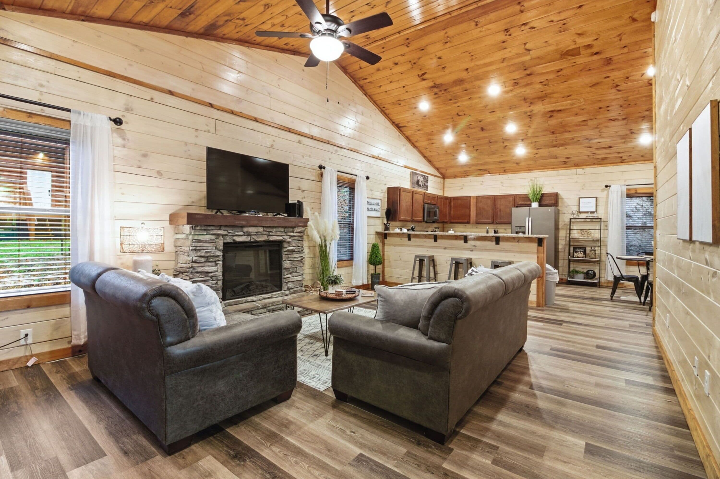 A log cabin interior with a stone fireplace, two grey sofas, a ceiling fan, and a kitchen area with wooden cabinets.