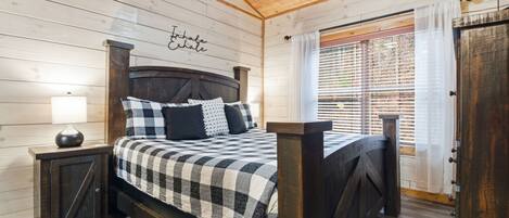 A bedroom with a wooden ceiling, a bed with a checkered bedspread, a nightstand with a lamp, and a window with blinds.
