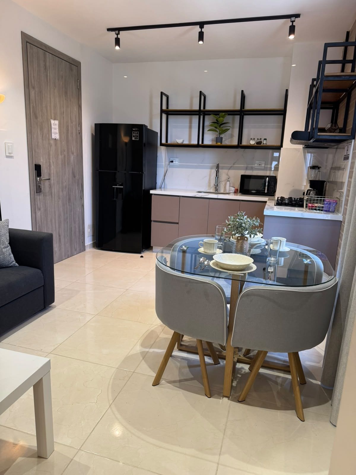 Uma sala de estar moderna com uma mesa de jantar de vidro, cadeiras cinzas, um sofá preto e uma cozinha com micro-ondas, fogão e armários.