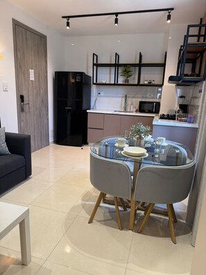Un salon moderne avec une table à manger en verre, des chaises grises, un canapé noir et un coin cuisine équipé d’un four à micro-ondes, d’une cuisinière et de placards.