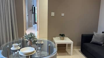 Una sala de estar moderna con una mesa de comedor de vidrio, dos sillas, un sofá negro, una mesita auxiliar blanca y un sofá gris.