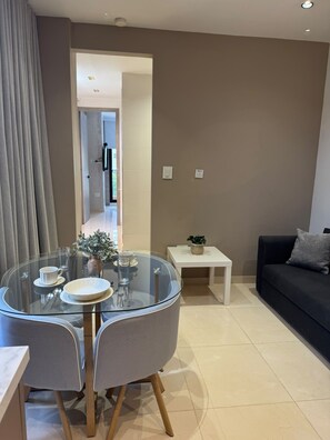 Un salon moderne avec une table à manger en verre, deux chaises, un canapé noir, une petite table d’appoint blanche et un divan gris.