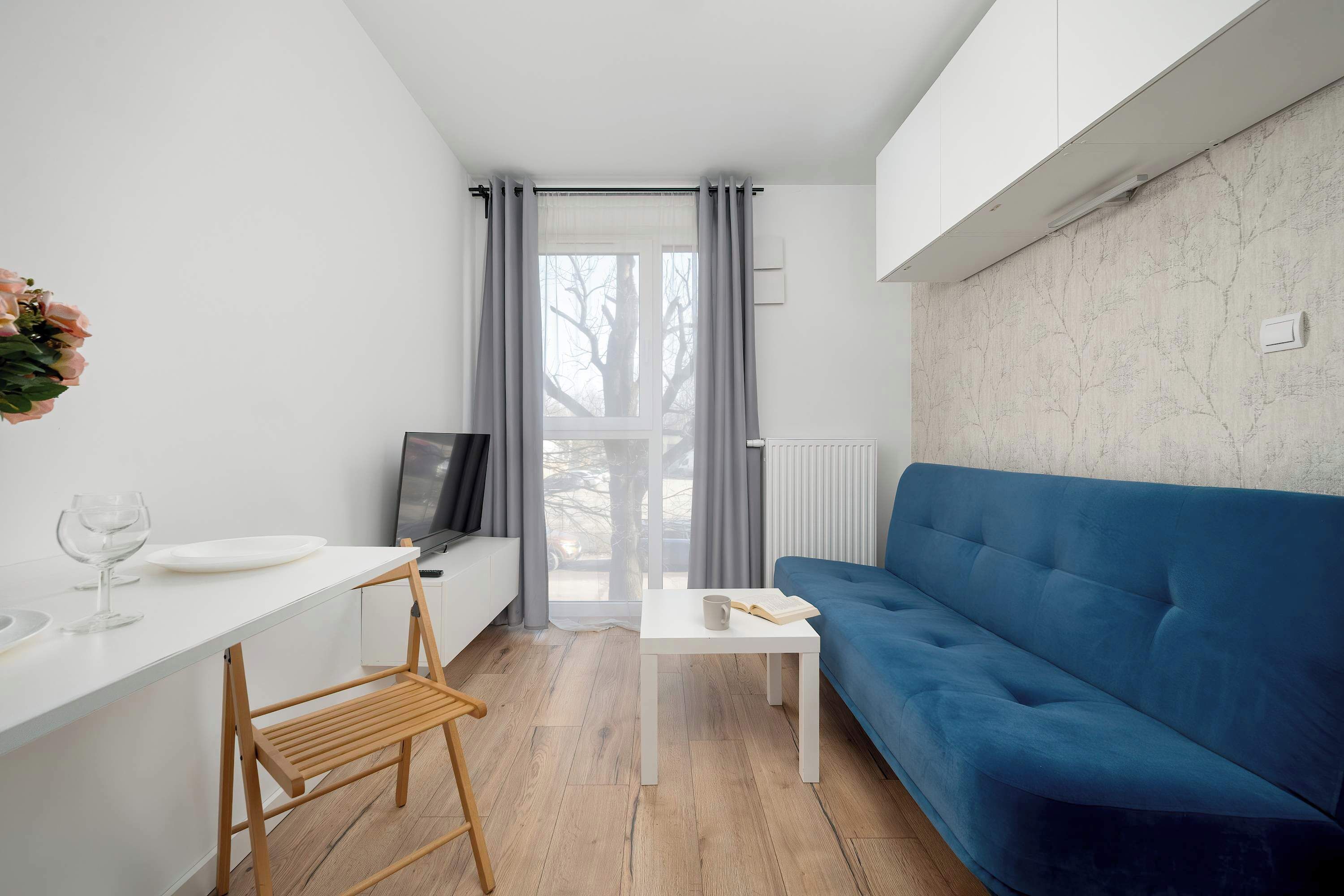 A bright living area with a blue sofa placed against the wall and a small white dining table with wooden chairs nearby. Large floor-to-ceiling windows allow natural light to fill the room. 