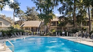 Une piscine dotée de chaises longues et d’un espace aménagé au bord de l’eau, le tout entouré d’arbres et de bâtiments.