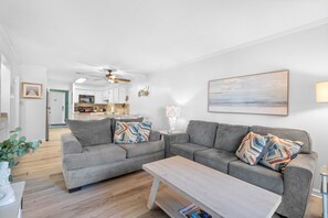 Un salon moderne avec un canapé gris, une table basse en bois et une cuisine en arrière-plan.