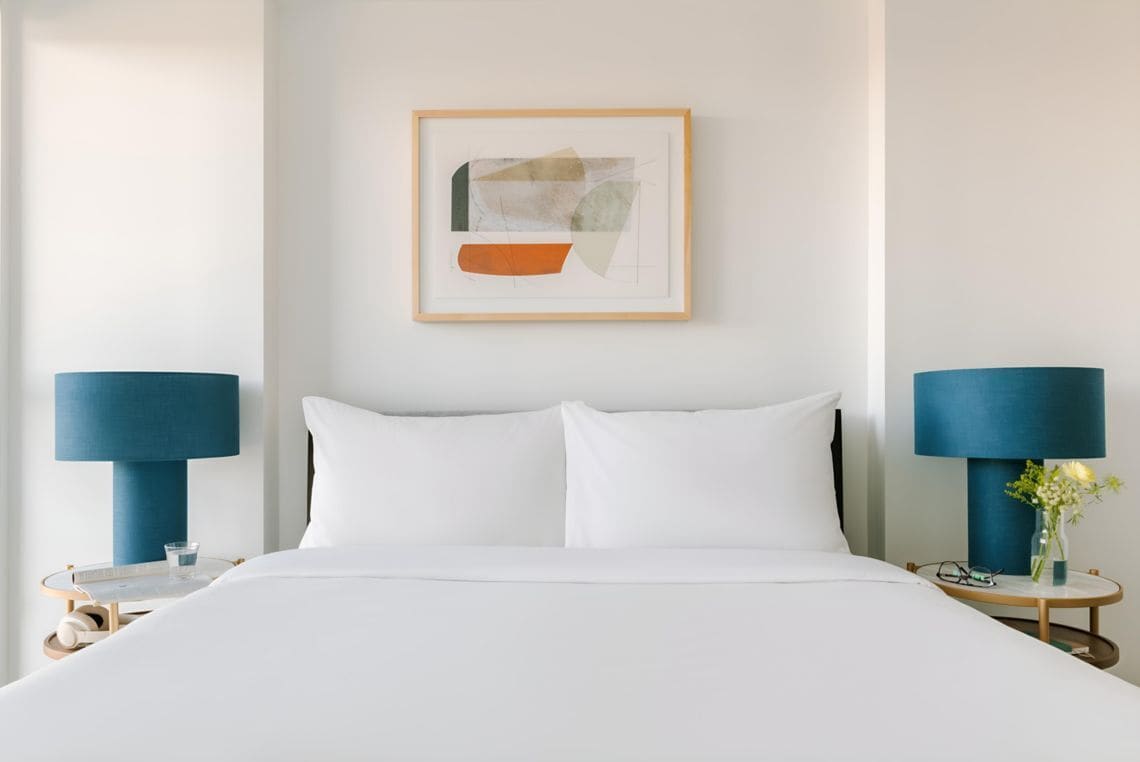 Habitación de hotel con cama, dos mesitas de noche con lámparas, una obra de arte abstracta enmarcada en la pared y un jarrón con flores sobre una de las mesitas.