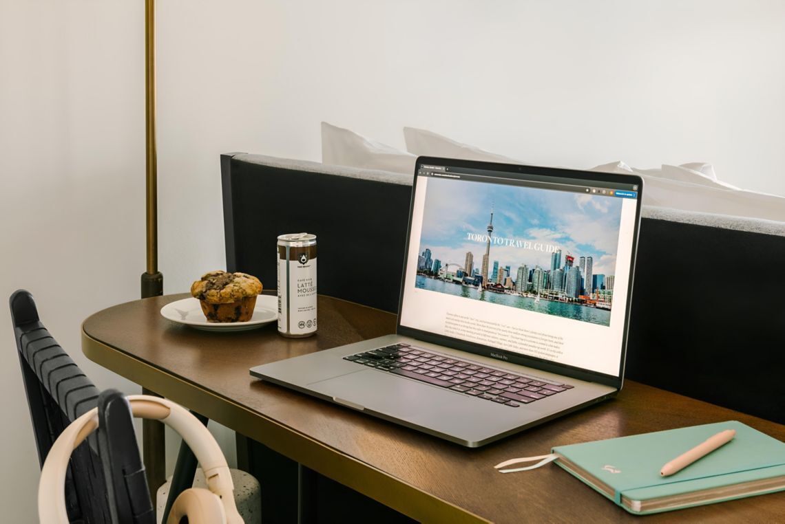 Een laptop op een houten tafel waarop een reisgids voor Toronto, een muffin, een koffiekop en een notitieboek liggen.