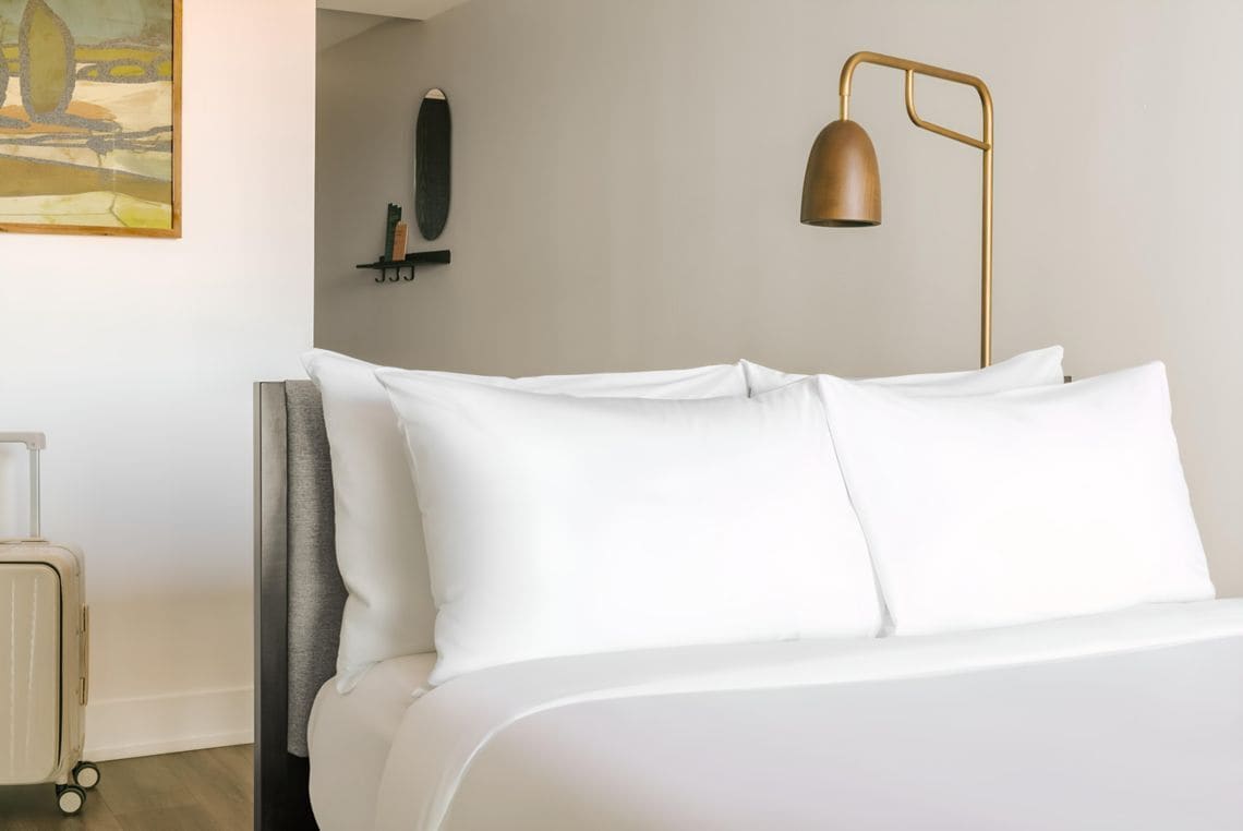 A neatly made bed with white linens, a gold floor lamp, and a painting on the wall.