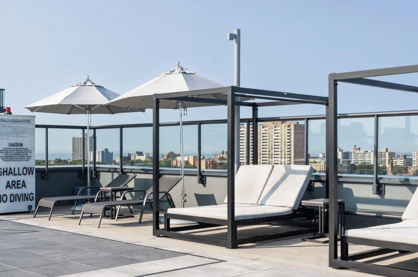 A rooftop lounge with sunbeds and umbrellas, a sign warning of shallow water, and a cityscape view.