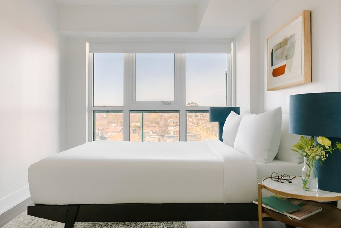 Un dormitorio con un ventanal grande, una cama, una mesita de noche con una lámpara y un cuadro enmarcado en la pared.