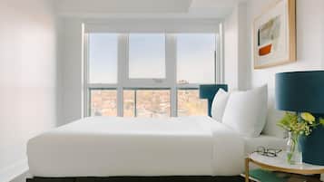 Un dormitorio con un ventanal grande, una cama, una mesita de noche con una lámpara y un cuadro enmarcado en la pared.