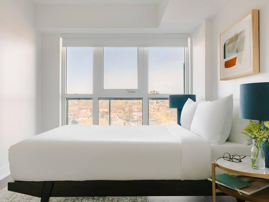 Un dormitorio con un ventanal grande, una cama, una mesita de noche con una lámpara y un cuadro enmarcado en la pared.