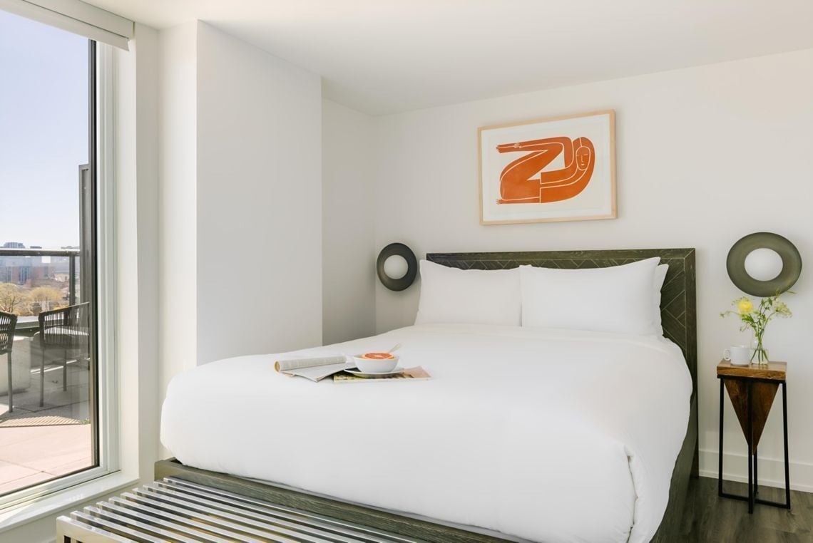 Ein Hotelzimmer mit einem großen Bett, einem Fenster mit Blick, einem gerahmten Kunstwerk an der Wand und einem kleinen Beistelltisch mit Blumen.