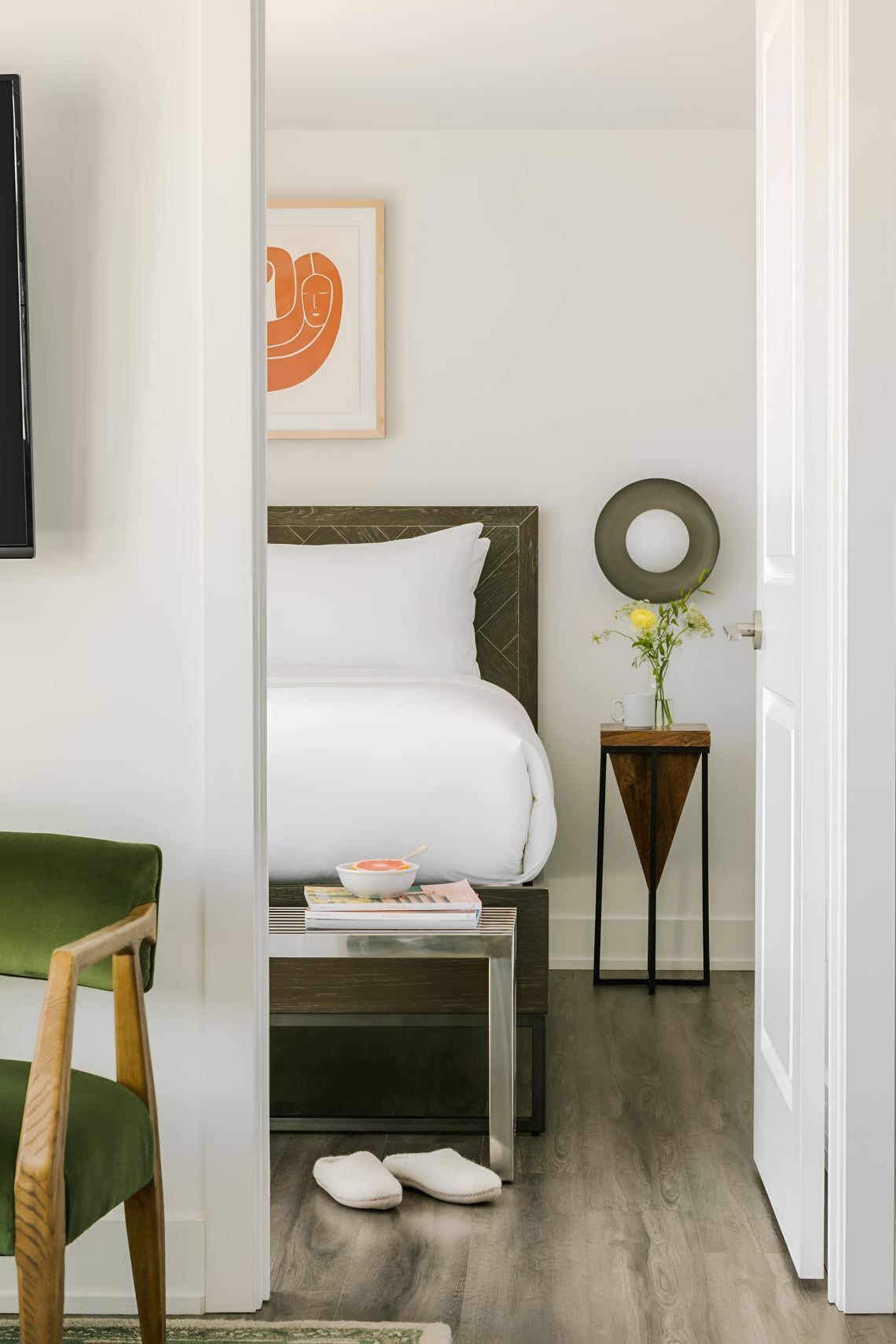 Una habitació d'hotel amb llit, taulella de nit, una taula petita amb un ram i una cadira verda, i xancletes a terra.