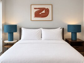 Een net opgemaakt bed met witte lakens, met aan beide zijden nachtkastjes met blauwe lampen, en een ingelijst kunstwerk erboven.
