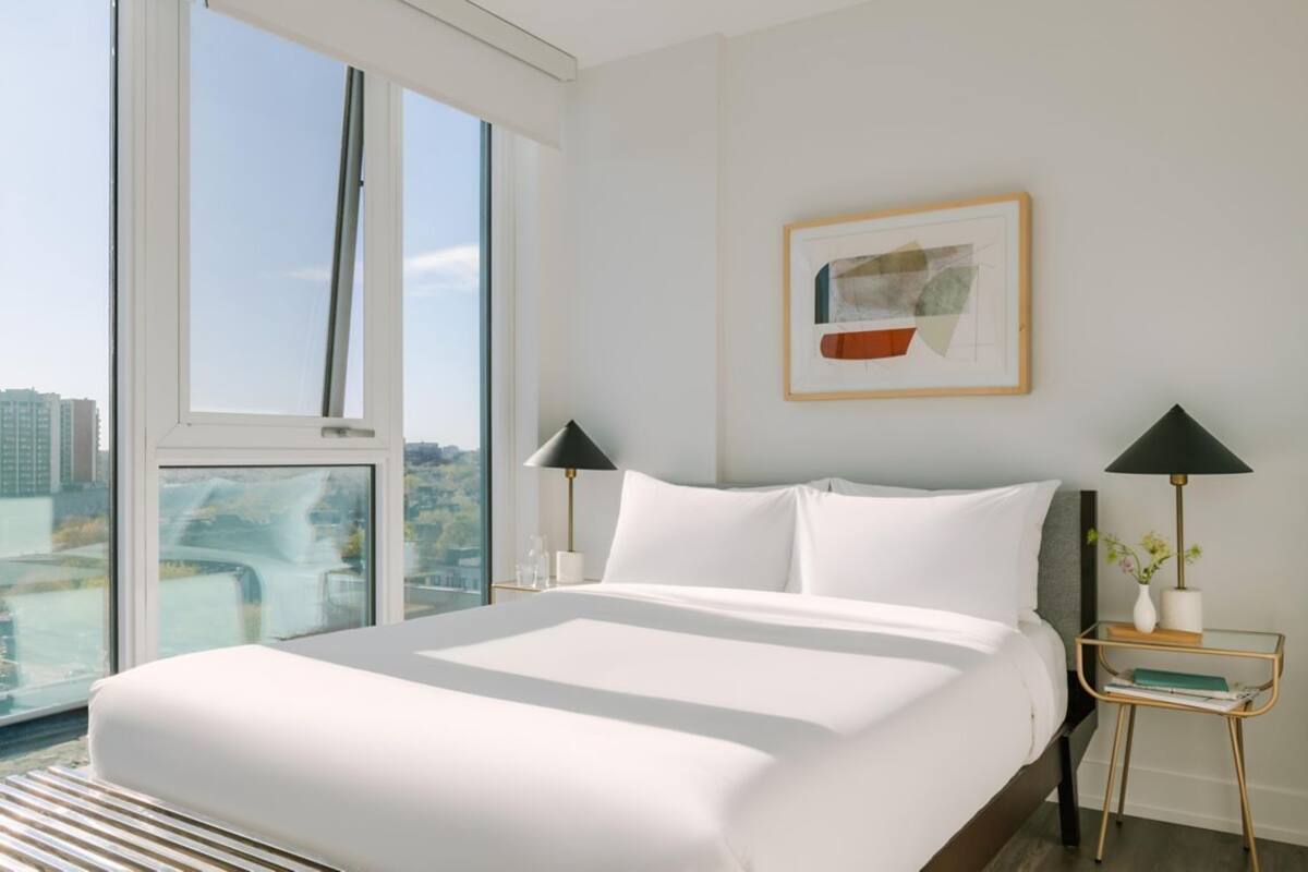 Une chambre d’hôtel avec un grand lit, deux tables de chevet avec des lampes, une œuvre d’art encadrée au mur et une vue sur la ville à travers de grandes fenêtres.