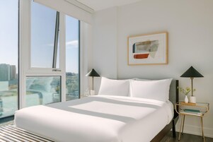 Hotelkamer met een groot bed, twee nachtkastjes met lampen, een kunstwerk in een lijst aan de muur en uitzicht op de stad door grote ramen.
