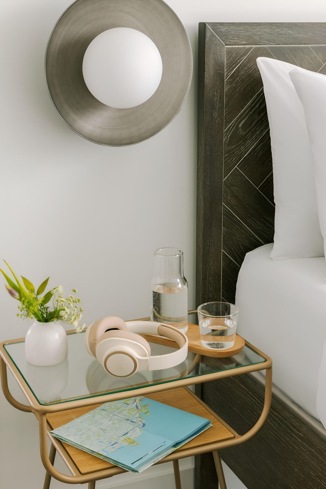 A neatly arranged bedside table with a map, a glass of water, a bottle, headphones, and a small plant.