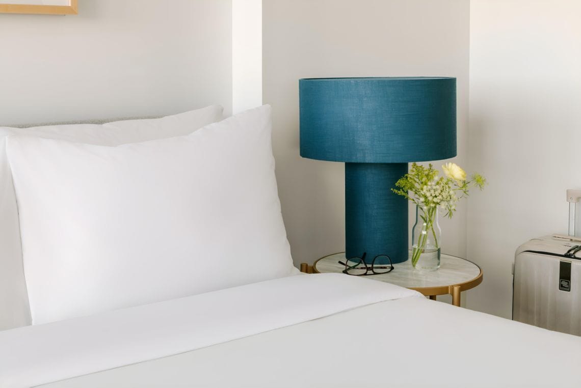 A hotel room with a bed, a blue lamp, a small table with a vase of flowers, and a suitcase.