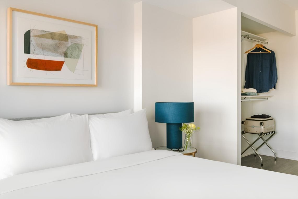A hotel room with a bed, a framed abstract painting, a bedside lamp, a small table with a vase, and a closet with hanging clothes.