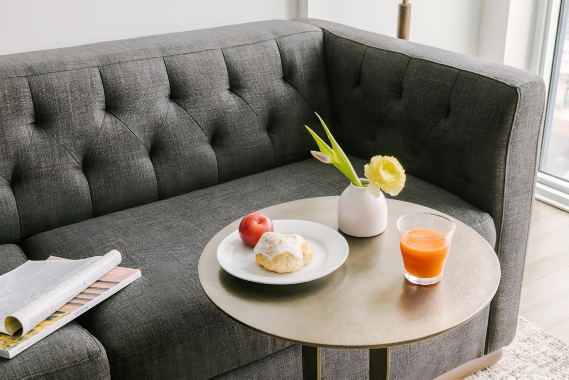 Um sofá cinza estofado com uma mesa redonda ao lado, exibindo um prato com um pastel, um copo de suco de laranja e um vaso com flores.