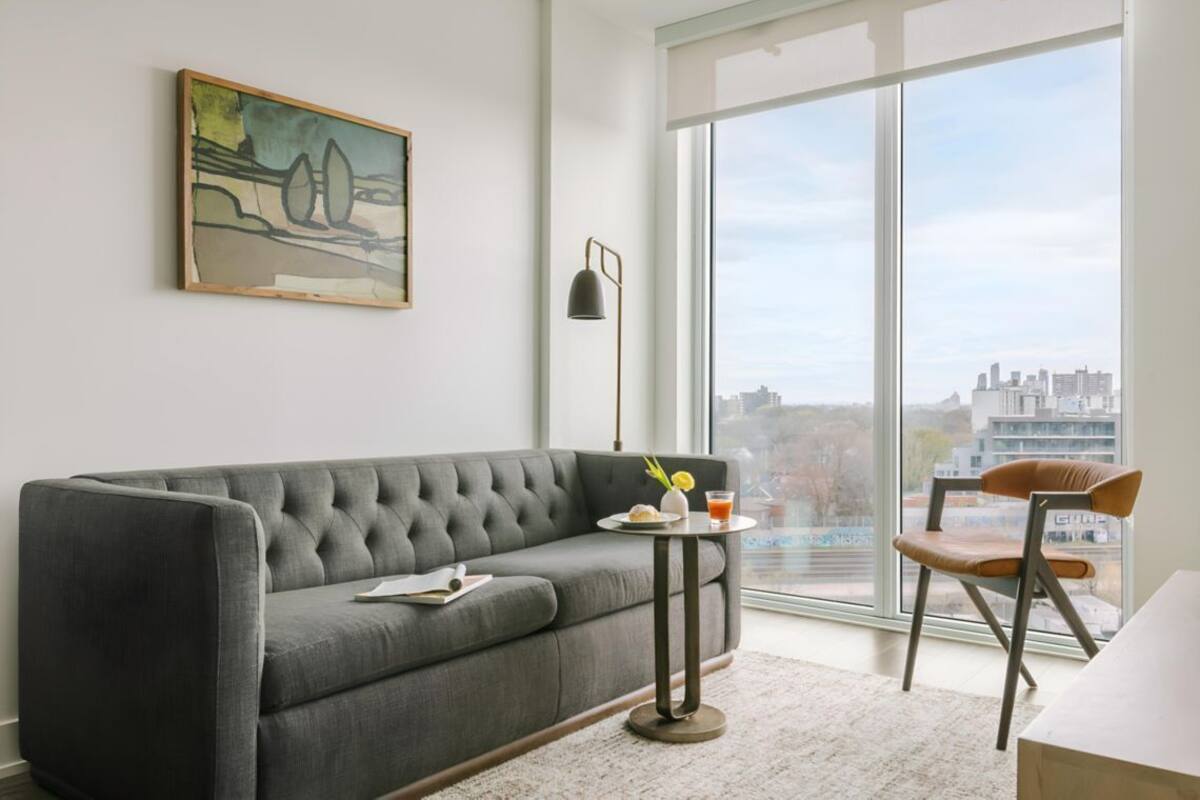 Un salon moderne avec un canapé gris, une petite table ronde sur laquelle se trouve une boisson, un lampadaire et un fauteuil. Une grande fenêtre offre une vue sur le paysage urbain.