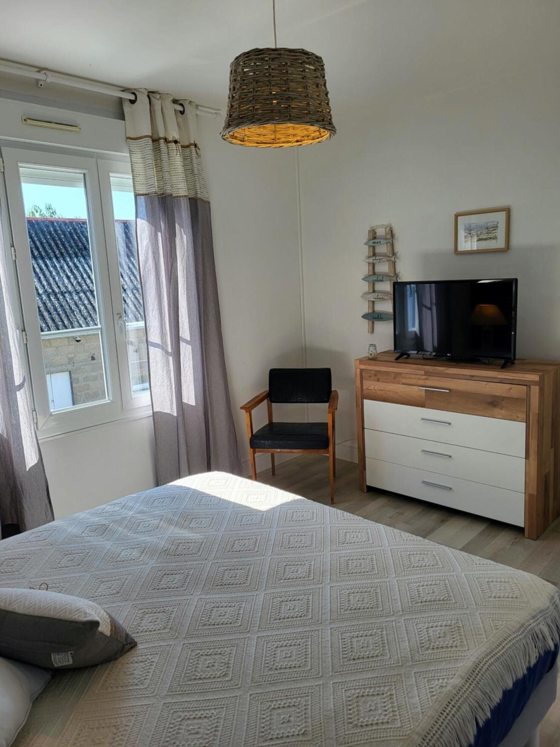 A bedroom with a bed, a chair, a TV on a wooden dresser, and a window with curtains.