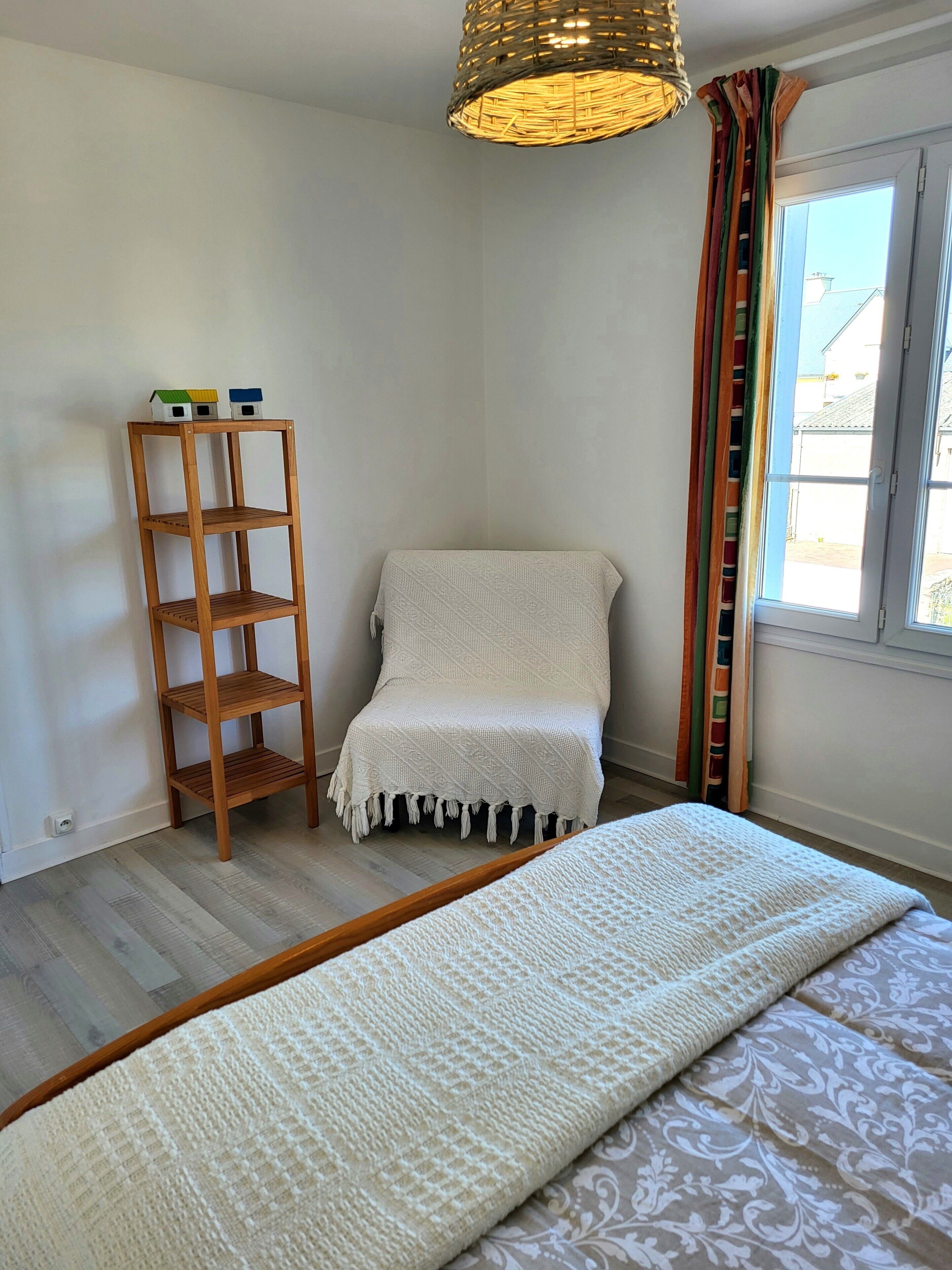 Een slaapkamer met een houten plank, een witte stoel, een bed met een witte deken en een raam met gordijnen.