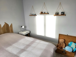 A bedroom with a bed, bedside table, lamp, hanging shelves, and a wooden chest with stuffed toys.