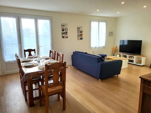 A dining area with a table set for four, a blue sofa, a TV on a wooden stand, and a wall with framed pictures.