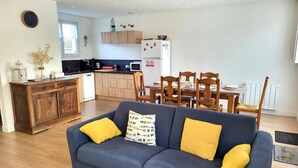 A modern living room with a sofa, wooden dining table, and kitchen area with cabinets and appliances. - Holiday Home "La Villa Kikifil, Seaside" with Private Terrace, Garden & Wi-Fi (Saint-Germain-sur-Ay)