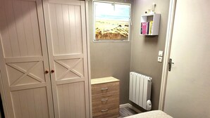 Ein Schlafzimmer mit Bett, Kleiderschrank, Schminktisch, Heizkörper und Fenster mit Blick auf ein Feld.