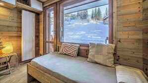 Una habitación cálida tipo cabaña con una cama, una mesita con una lámpara y un ventanal con vistas a una montaña nevada.