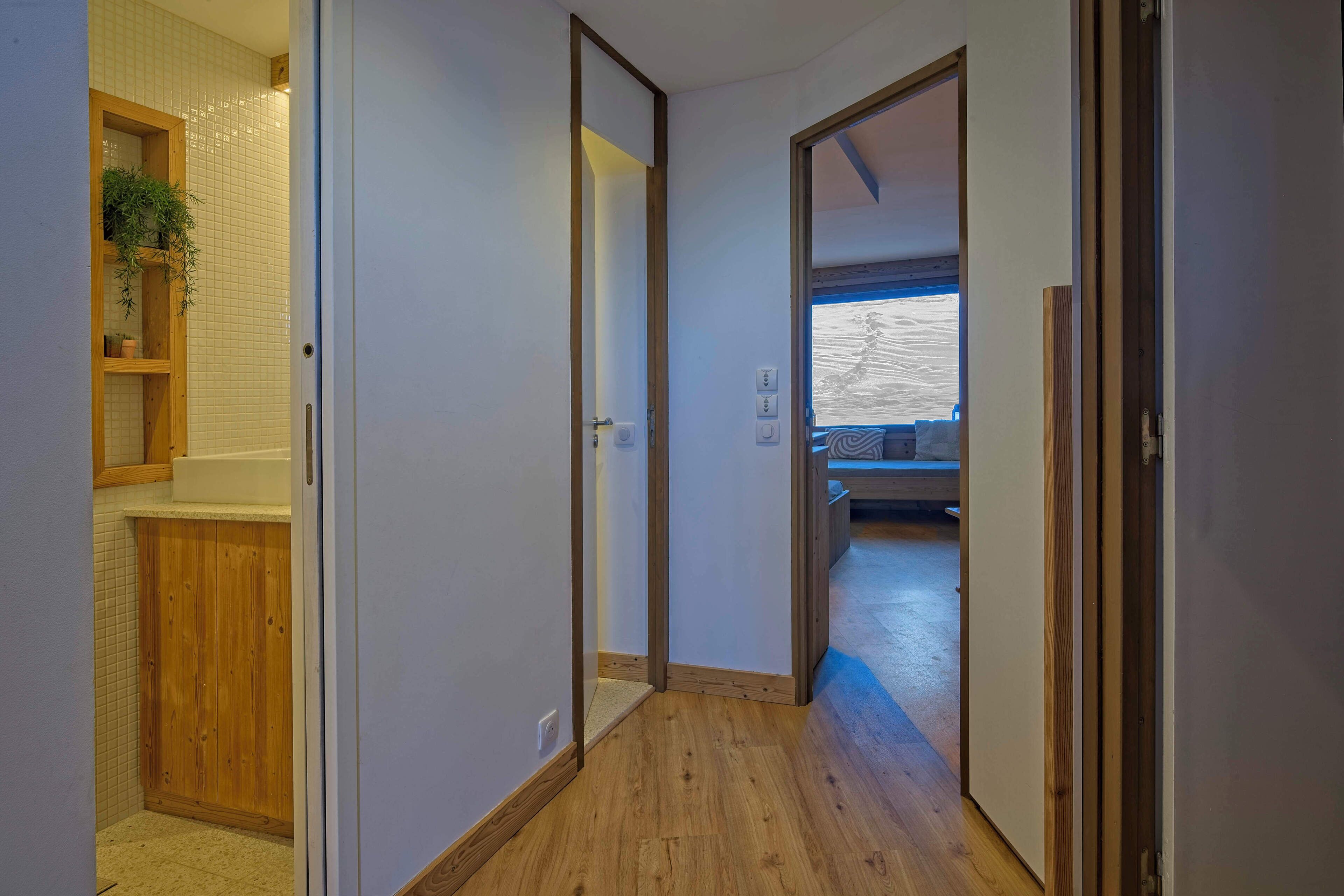 A hallway with wooden flooring, a wooden cabinet with a plant, and a view into a room with a sofa and a window.