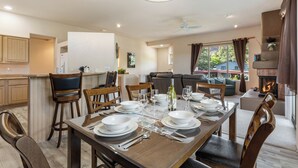 Un comedor con mesa de madera dispuesta para cuatro personas, una cocina al fondo y una sala de estar con chimenea.