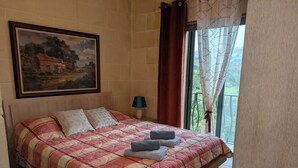 Un dormitorio con cama, almohadas, un cuadro en la pared, una mesita de noche, una lámpara y vistas al verdor a través de la ventana.